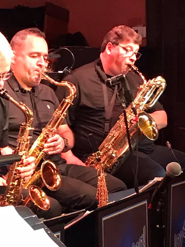 Two male musicians playing saxophones in a dark, red-lit venue