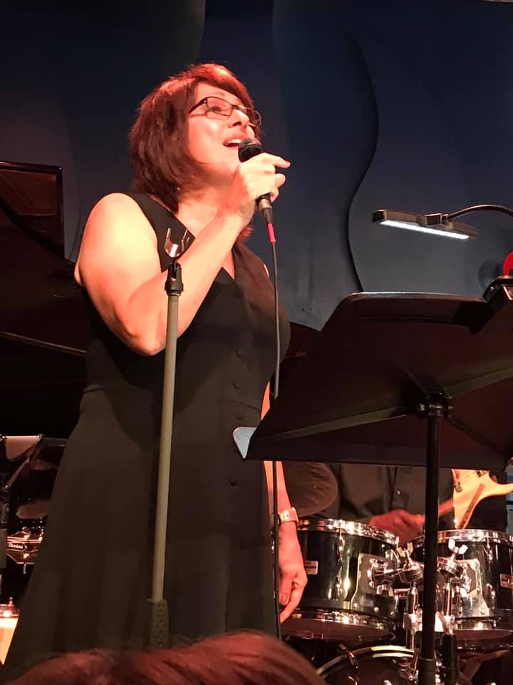 Woman with auburn hair and glasses singing into a microphone on stage