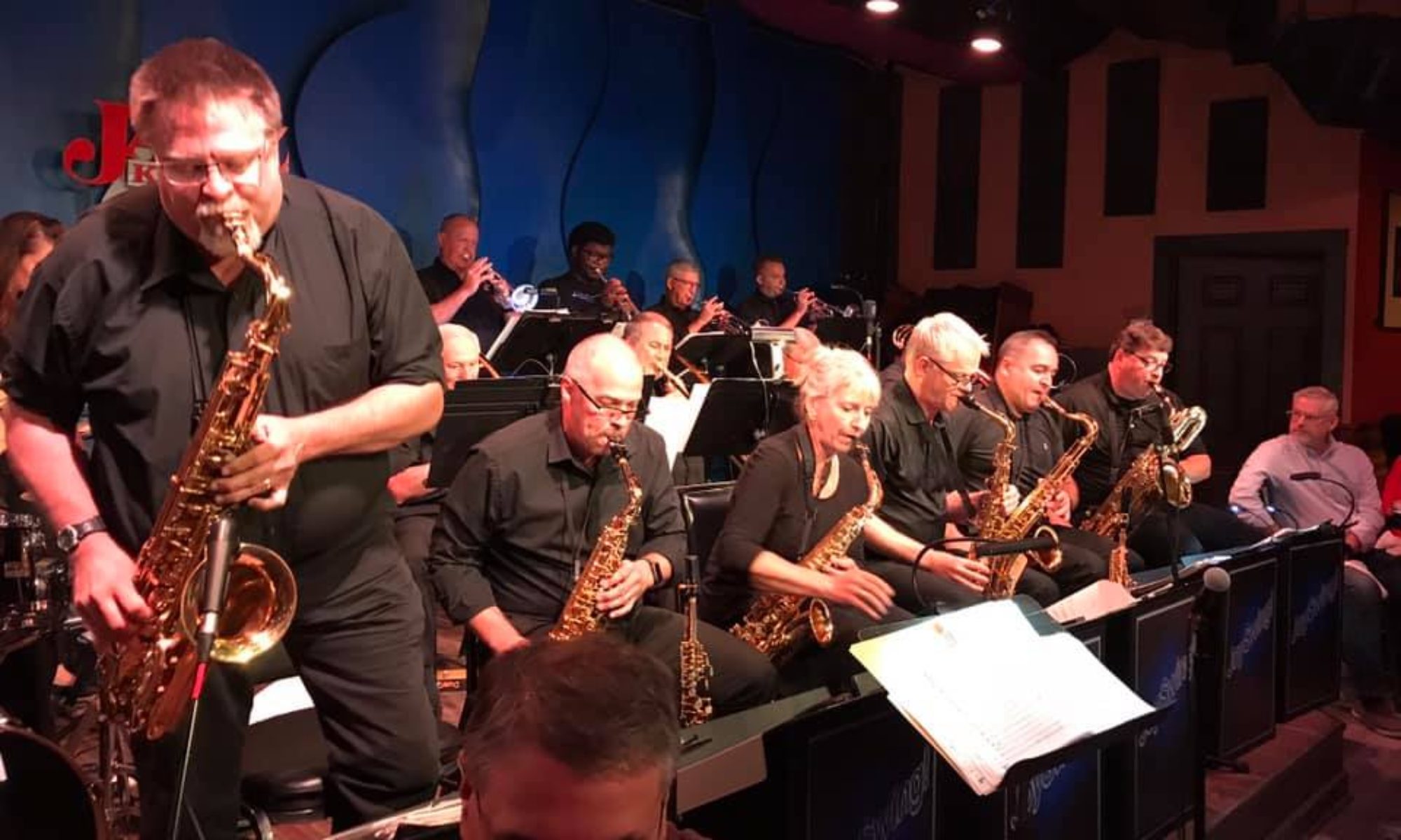 Large jazz orchestra playing their instruments on a dimly lit stage at The Jazz Kitchen